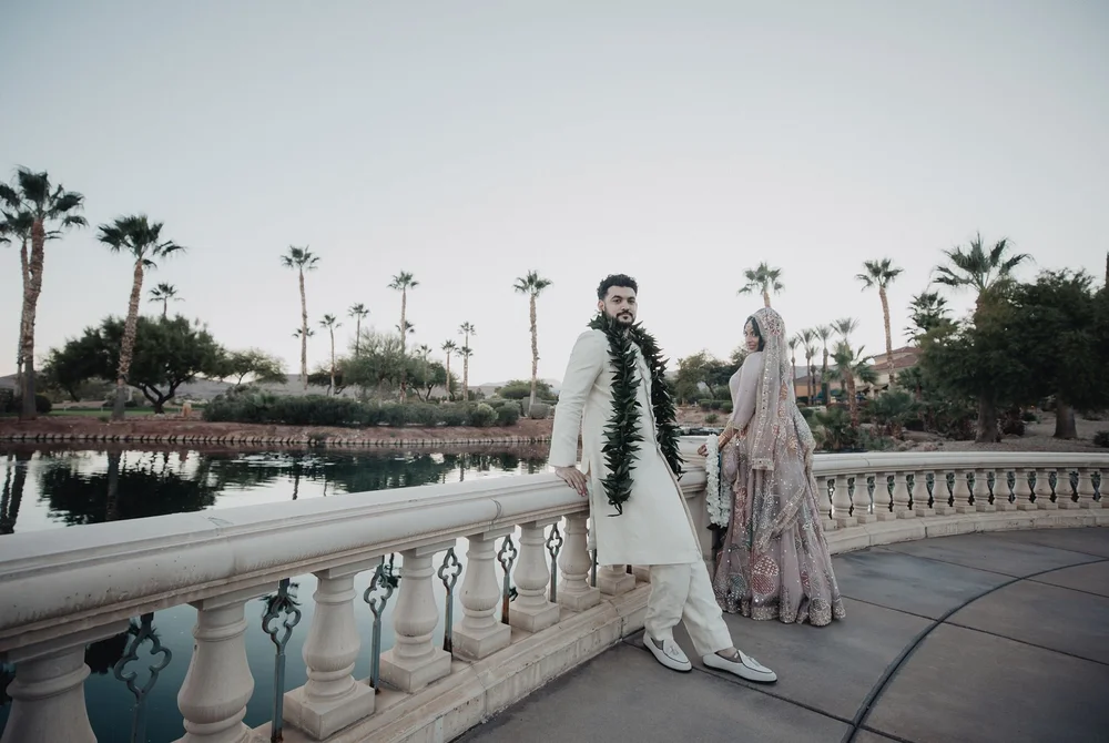 Groom in an ivory sherwani and garland beside the bride on a balustraded terrace by a lake