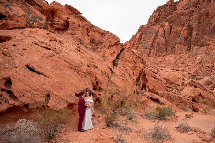 Groom in a burgundy suit and veiled bride stand close together in a hollow of wind-carved red sandstone at Valley of Fire.