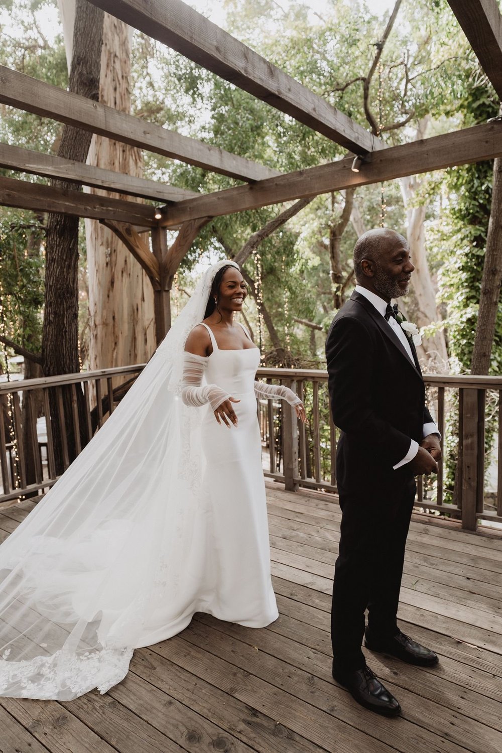 Bride in a fit-and-flare gown with lace sleeves and matching gloves standing under a wooden pergola with her father