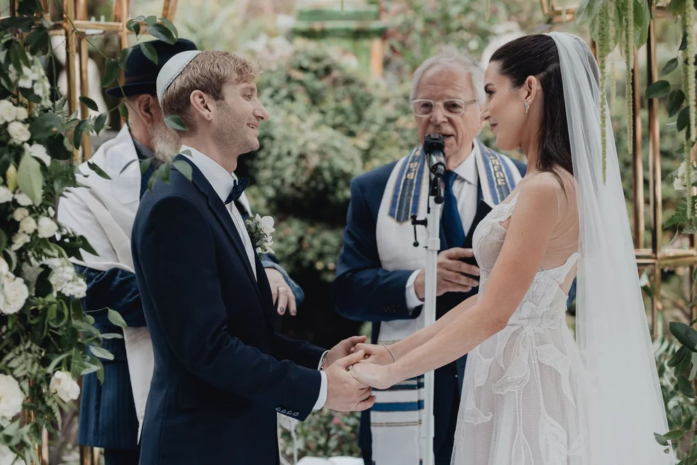 Rabbi speaking as the couple hold hands beneath a floral chuppah
