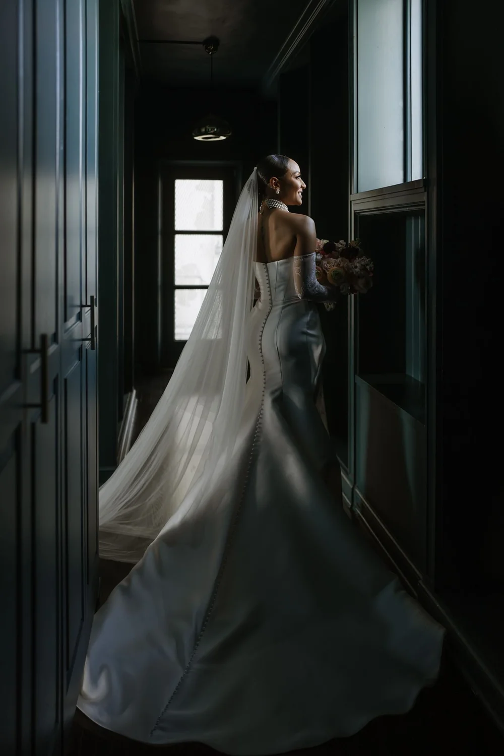 Back view of a bride in a fitted gown with a long lace-edged veil trailing behind her through a dark doorway