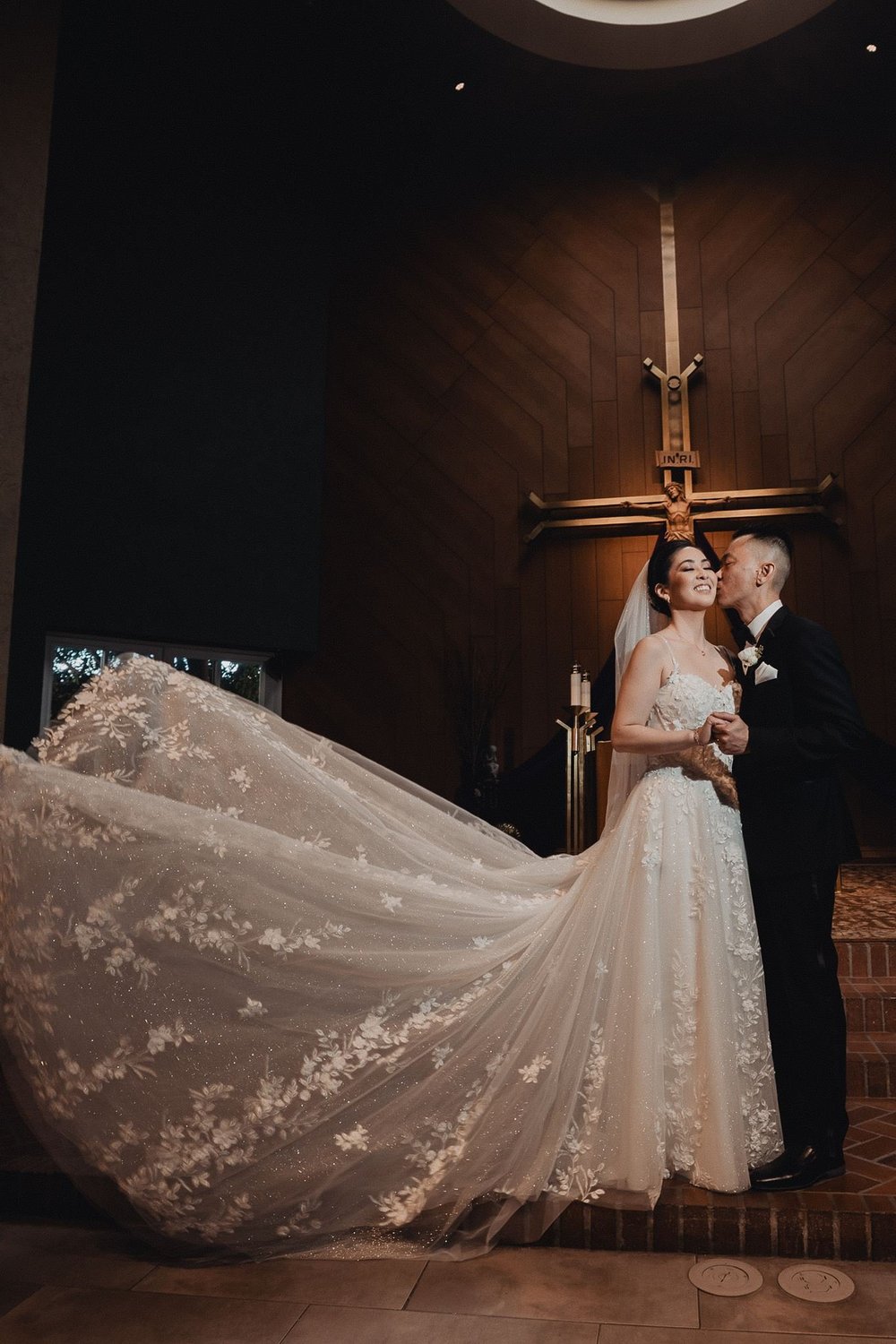 Bride in a fit-and-flare gown wearing a three-dimensional floral cathedral veil that sweeps across the floor beside her husband