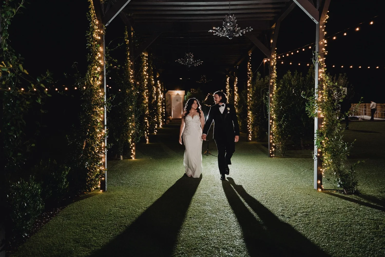 Couple walking hand in hand at night beneath a lit pergola wrapped in greenery and chandeliers at Terra in Murrieta