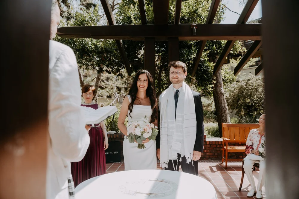 Couple standing behind the ketubah table under a wooden pergola, the groom wearing a tallit