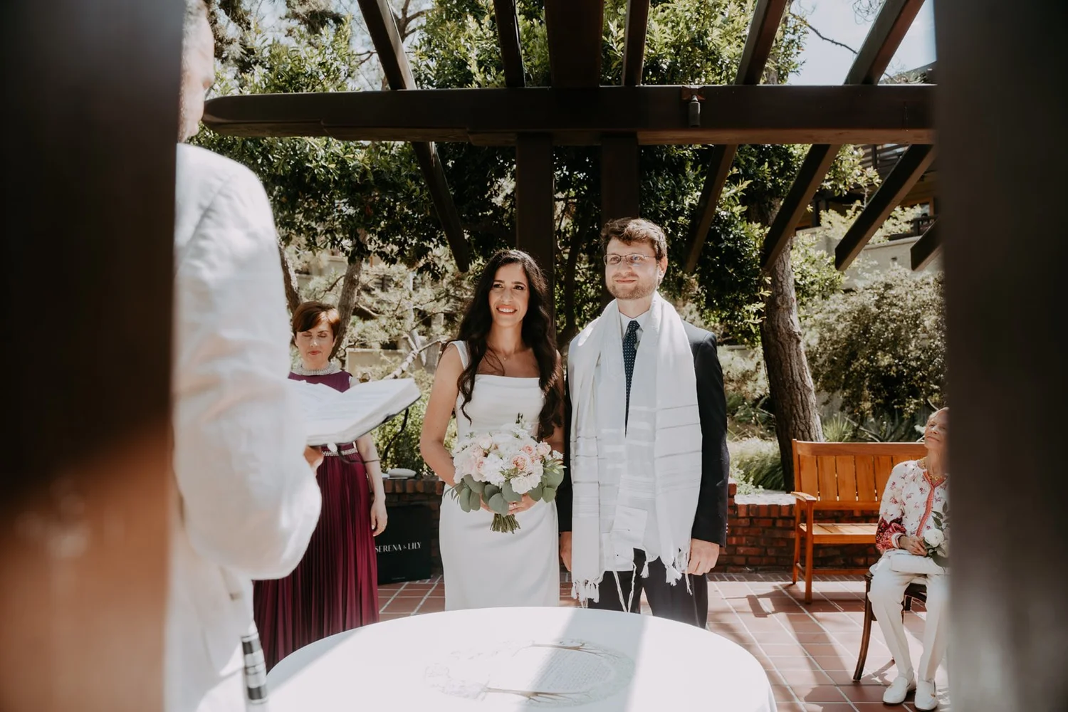Couple standing behind the ketubah table under a wooden pergola, the groom wearing a tallit
