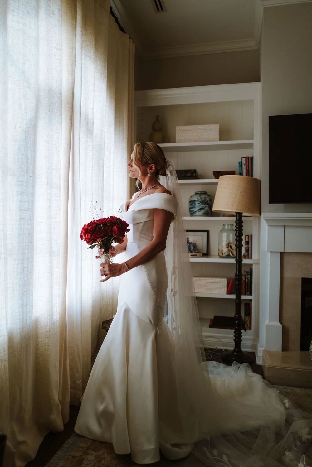 Bride in an off-the-shoulder white silk gown holding deep red roses in soft window light