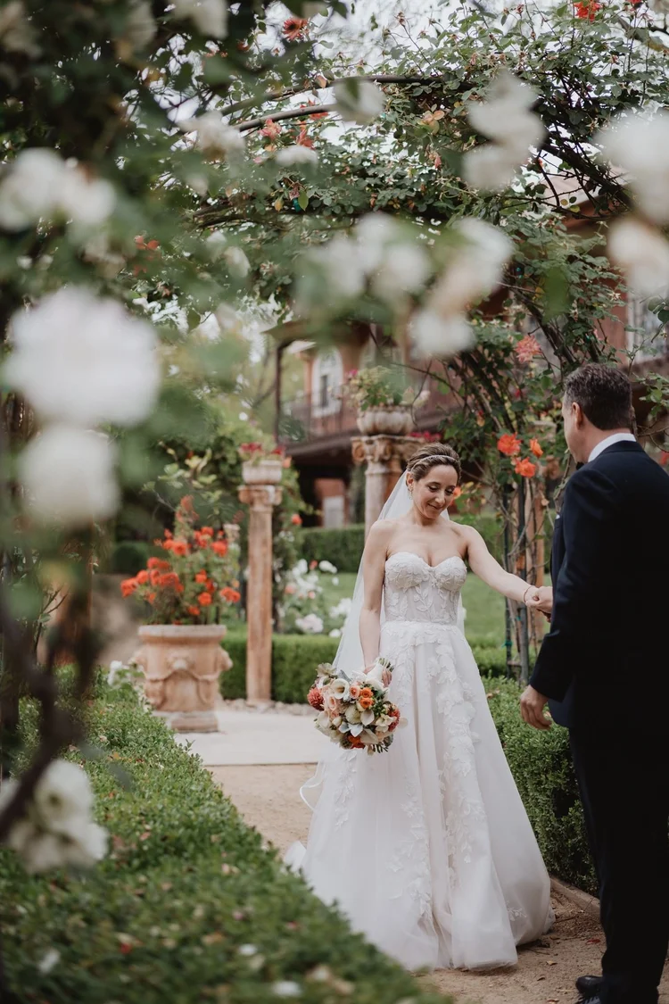 Bride in a lace A-line gown standing among flowering branches in a garden with her husband