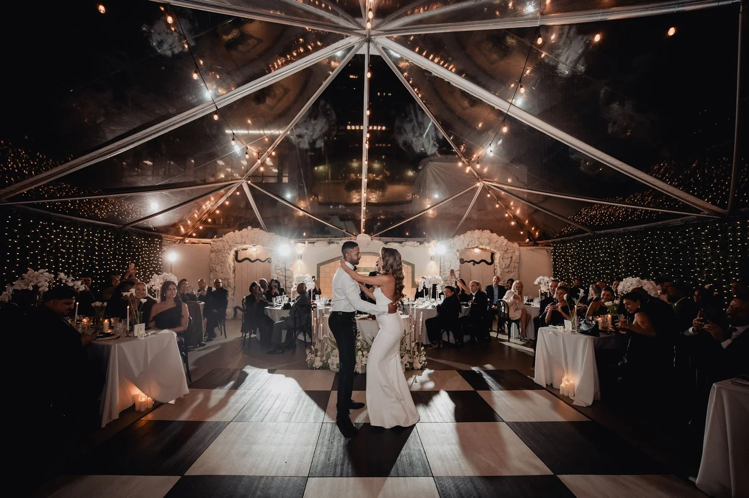 First dance under a clear-top tent on the Oviatt Penthouse terrace, guests seated around a black and white checkerboard floor