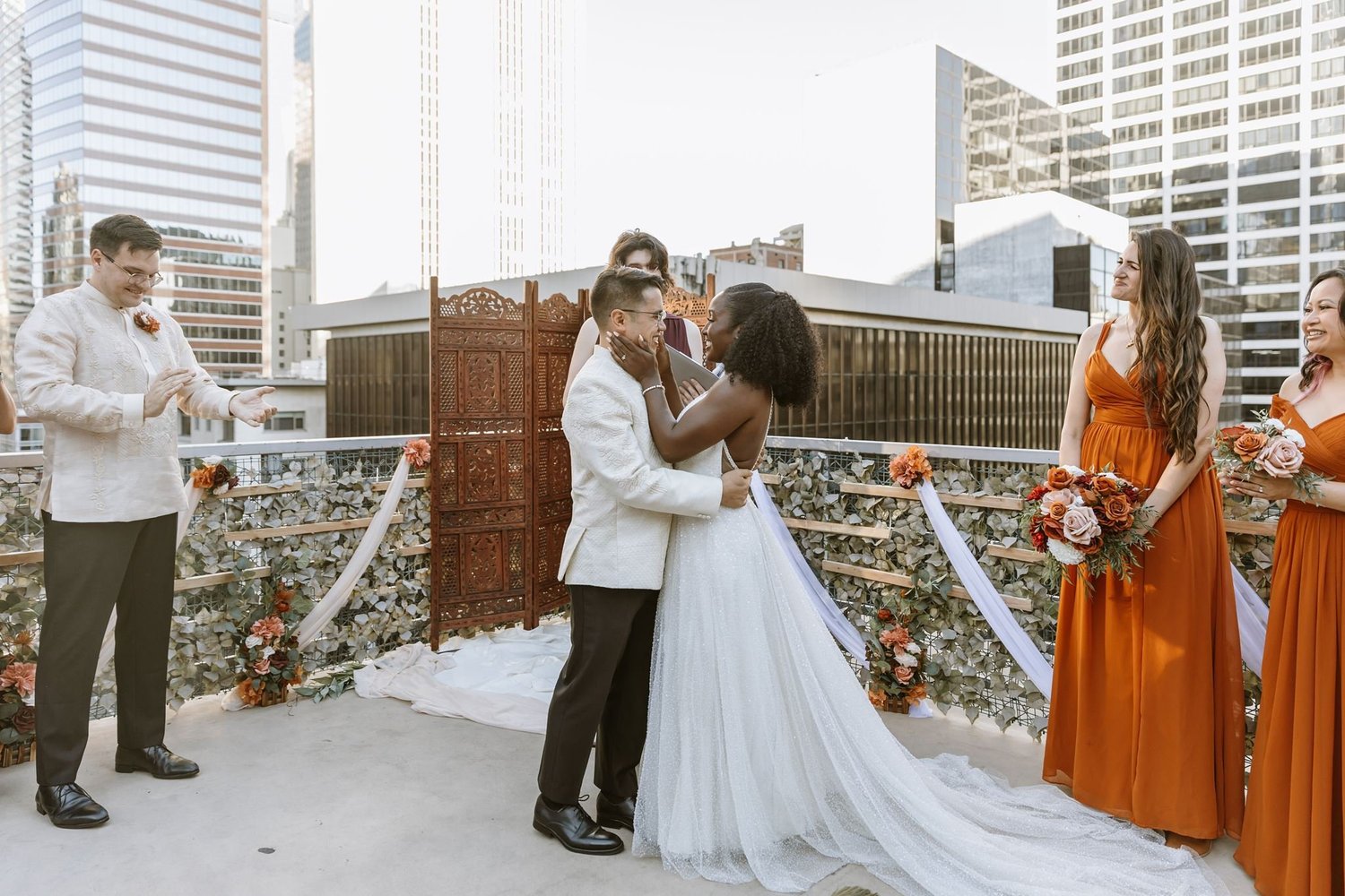 Bride and groom holding one another at the end of a rooftop ceremony at The Top of the Hub, bridesmaids in rust and downtown towers behind them