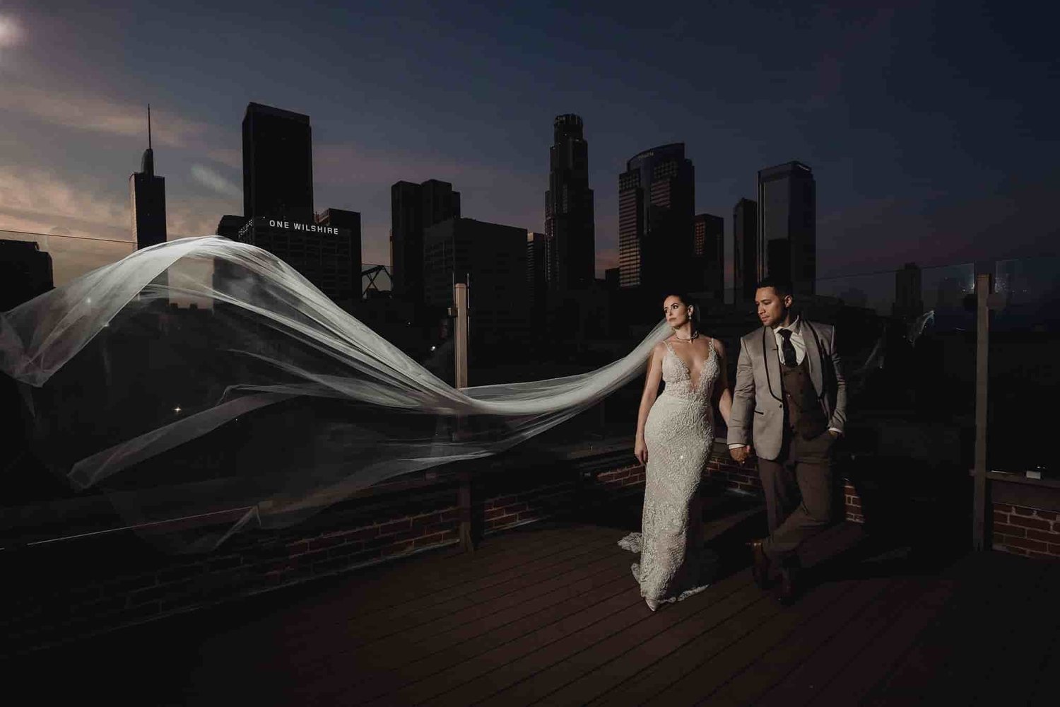 Bride in a fitted lace gown with an extremely long veil sweeping out in the wind against the downtown Los Angeles skyline at dusk