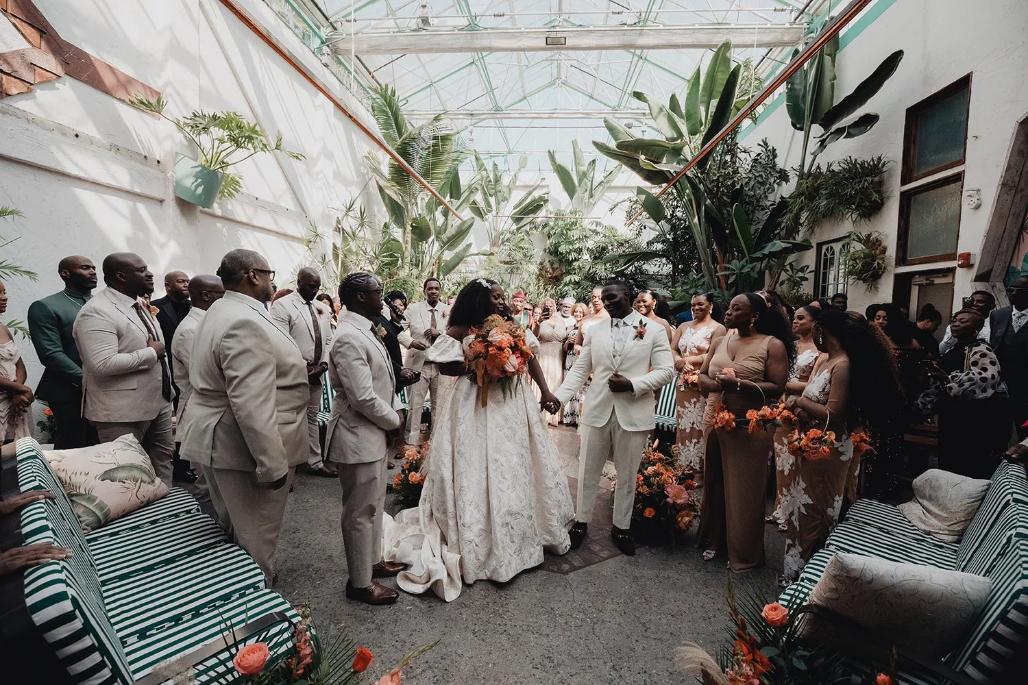 Wedding ceremony beneath the glass greenhouse roof at Valentine DTLA, guests seated on striped daybeds among tropical plants