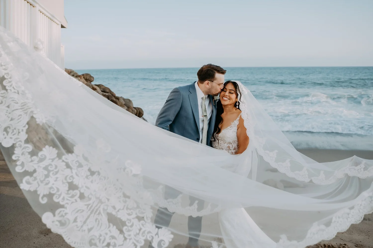 Long lace veil sweeping across the rocks as the couple stands above the ocean at Duke's Malibu