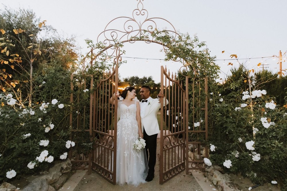 Couple framed by an ornate white gate covered in climbing white roses