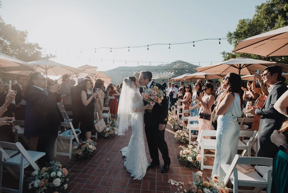 Couple kissing at the end of the aisle as guests applaud on the Castaway terrace under string lights