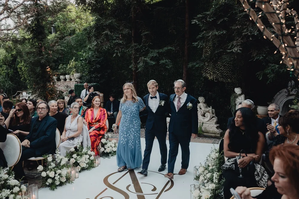 Parents walking the groom down a garden aisle to the chuppah