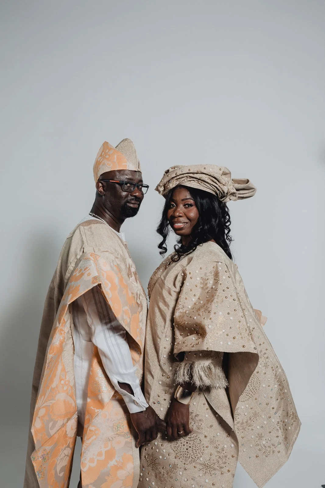 Nigerian couple in matching cream and peach aso ebi holding hands, the groom in an agbada and fila, the bride in a beaded gele
