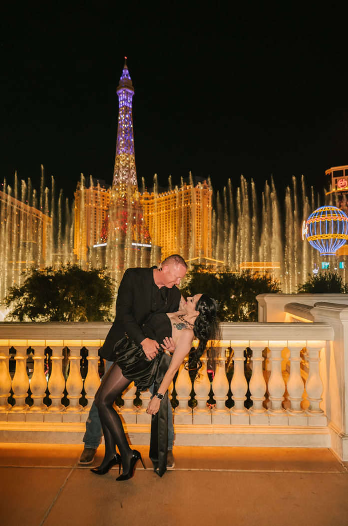 Man in a black blazer dips a woman in a black satin dress in front of the Bellagio fountains, with the Paris Las Vegas Eiffel Tower lit up at night.