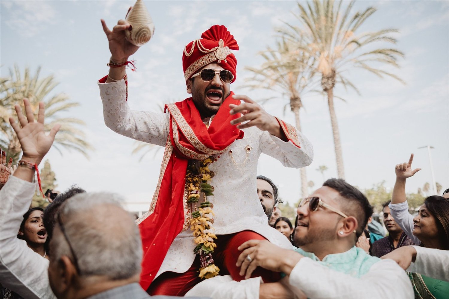 Groom in a cream sherwani and red safa lifted on shoulders during the baraat at the Hyatt Regency Huntington Beach, palms and blue sky behind