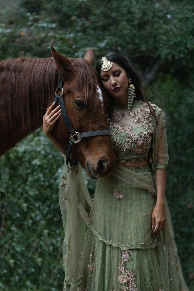 Bride in an emerald and gold lehenga resting her head against a horse before the ceremony