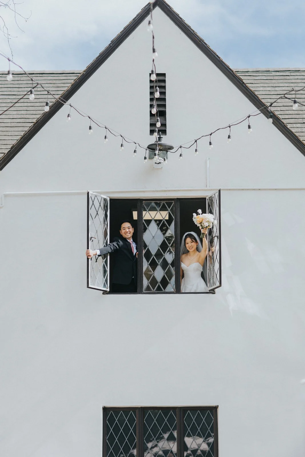Couple waving from a leaded-glass window in the white gable of The Sanctuary under string lights