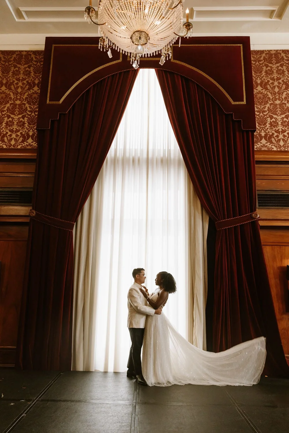Couple in an ornate lounge with heavy burgundy drapes, damask walls and a crystal chandelier overhead