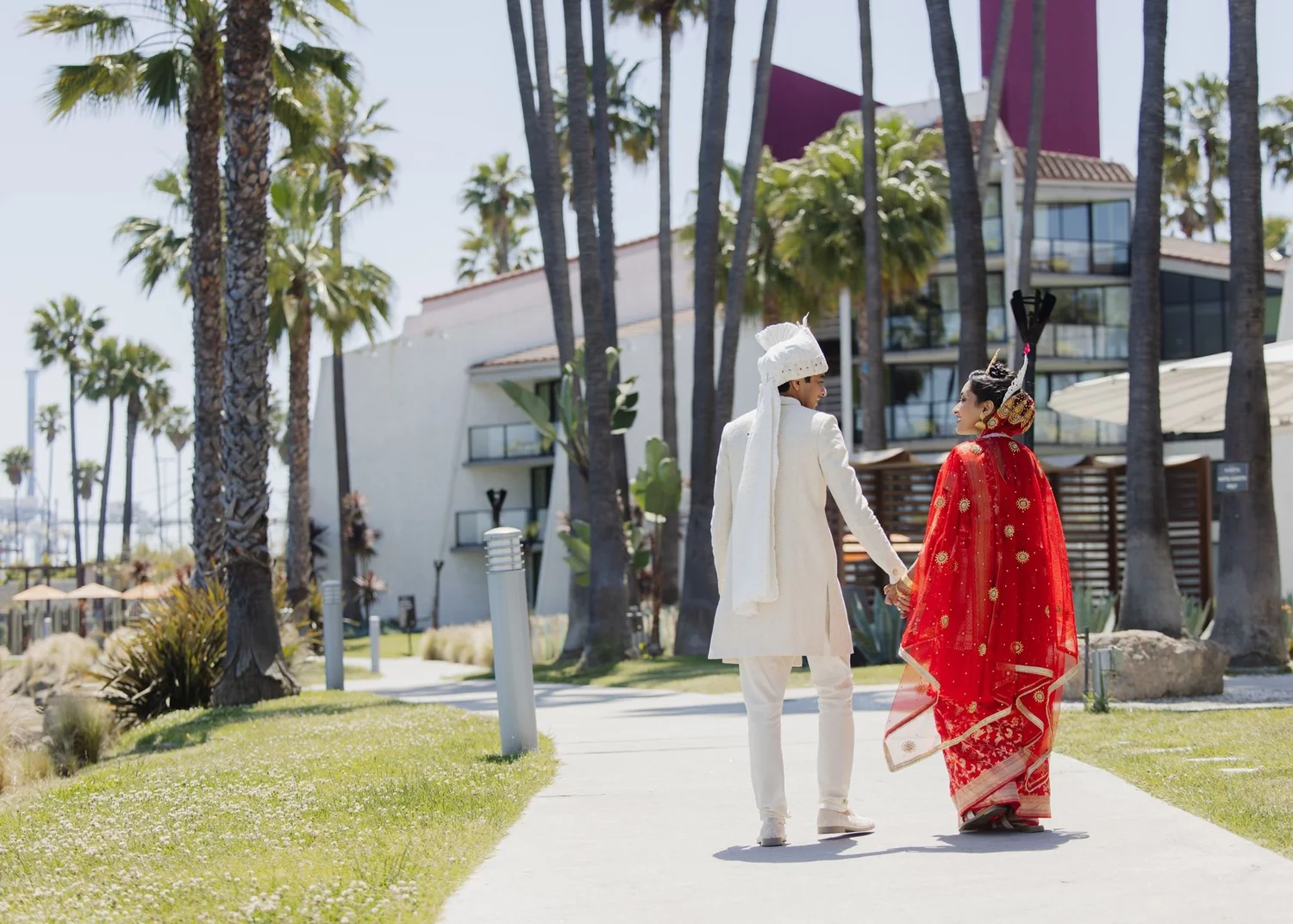 Couple in traditional South Asian wedding attire walking the palm-lined lawn at Hotel Maya