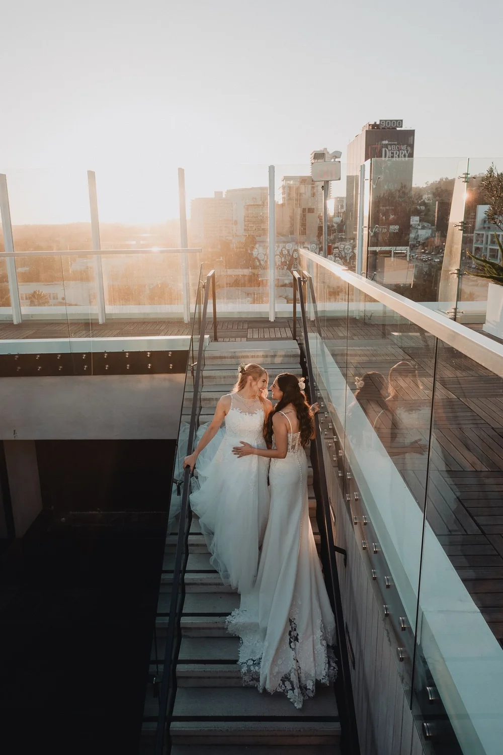 Two brides on the glass rooftop staircase at The London West Hollywood in low afternoon sun