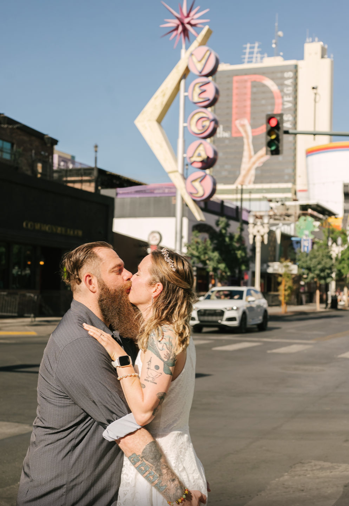 Bearded groom kisses his bride beneath the vintage VEGAS sign on Fremont Street in daylight, The D Las Vegas rising behind them.