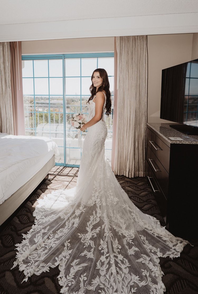 Bride in a lace mermaid gown with a three-dimensional floral sweep train beside a bright hotel window