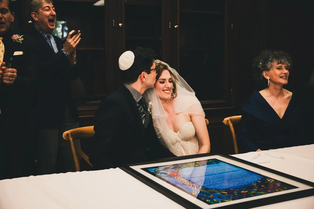 Couple leaning together and laughing over their signed ketubah as guests watch