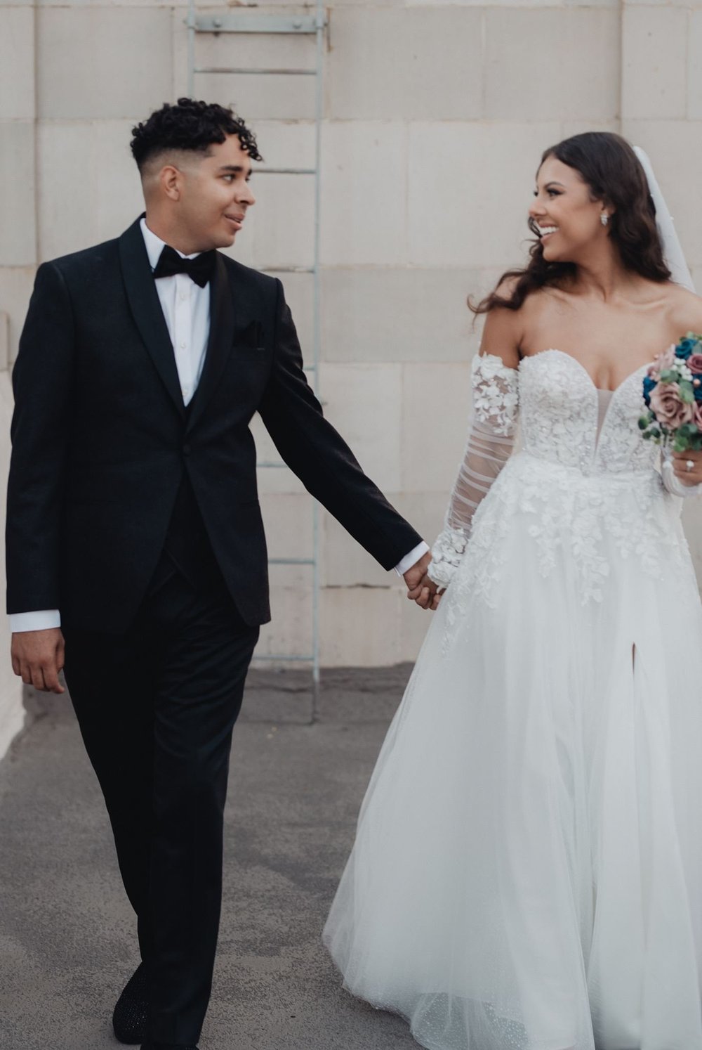 Bride in an A-line gown with sheer illusion lace long sleeves holding hands with her husband against a pale stone wall