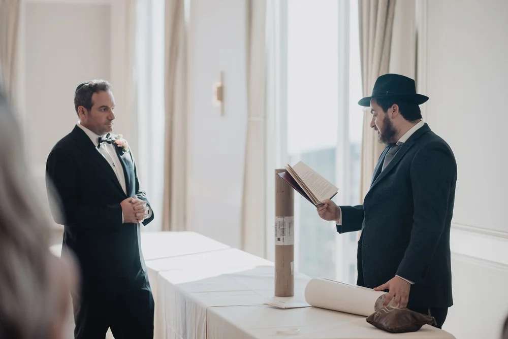 Rabbi in a black hat reading the ketubah aloud to the groom beside the signing table