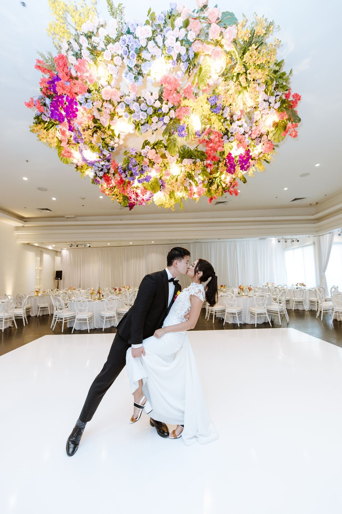 Groom dipping the bride under a large floral chandelier in the white room at Venue by Three Petals