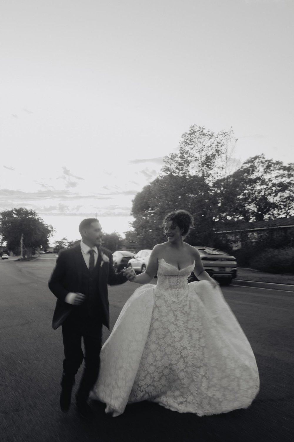 Black and white frame of a bride in a 3D floral ball gown holding the skirt as she crosses a road with her father