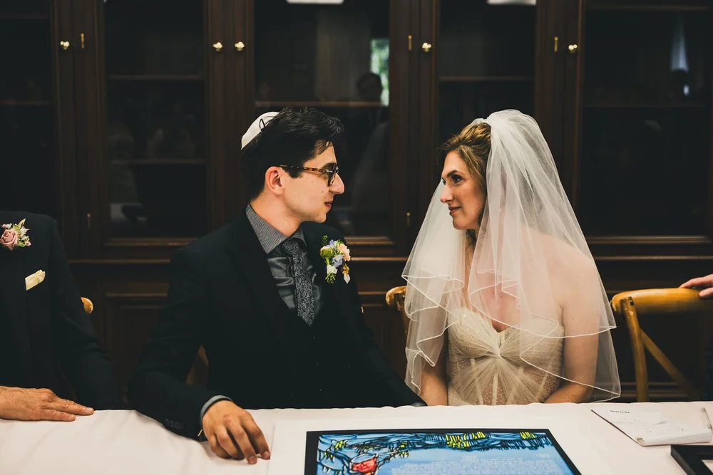 Bride and groom seated on opposite sides of the ketubah looking at each other before signing