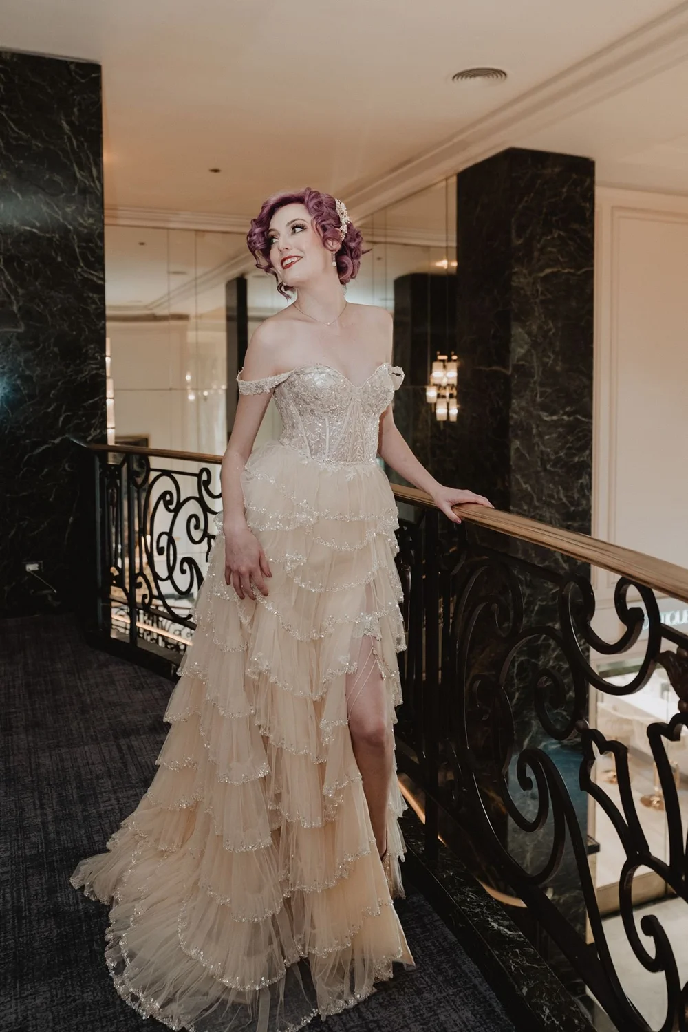 Bride in a blush pink ruffled gown with a high slit standing at an ornate iron staircase in a hotel lobby