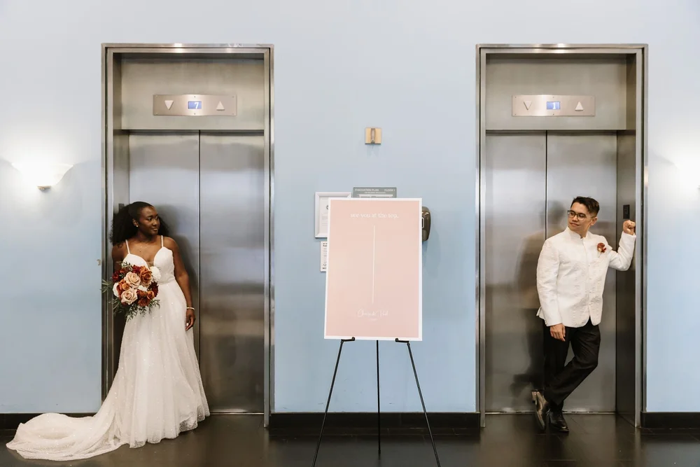 Bride and groom leaning against opposite lift doors beside a blush welcome sign on an easel