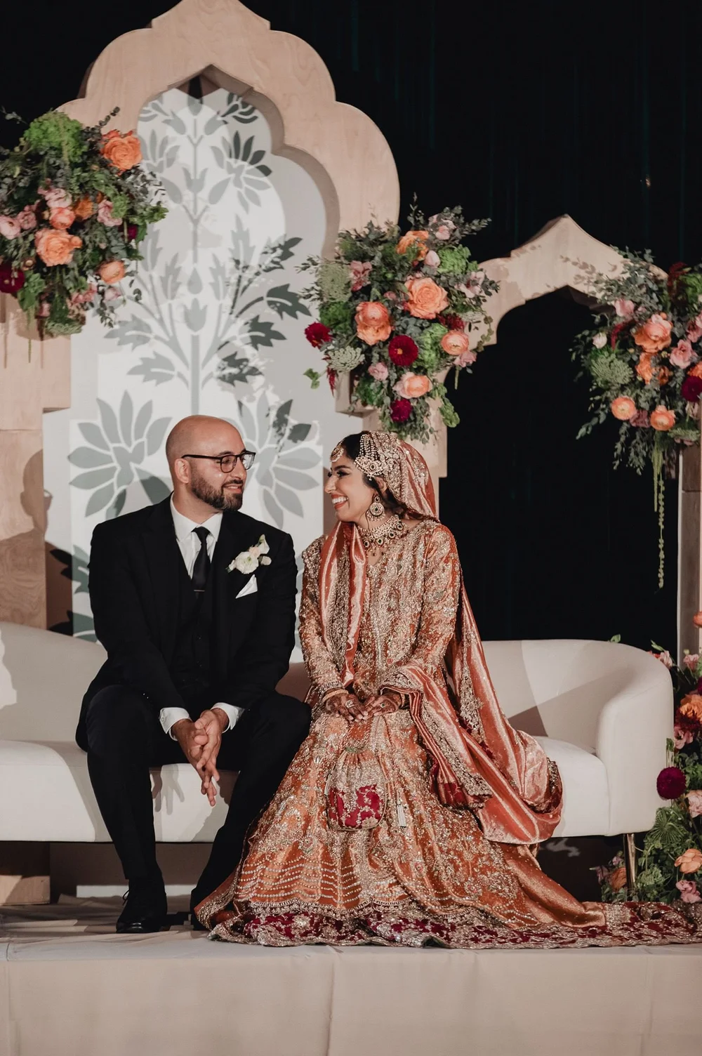 Bride and groom seated on the stage beneath a floral arch at a Muslim wedding reception