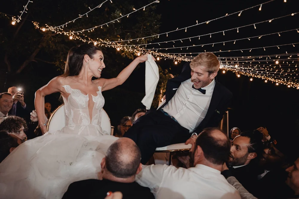 Bride and groom raised on chairs holding a napkin between them during the hora