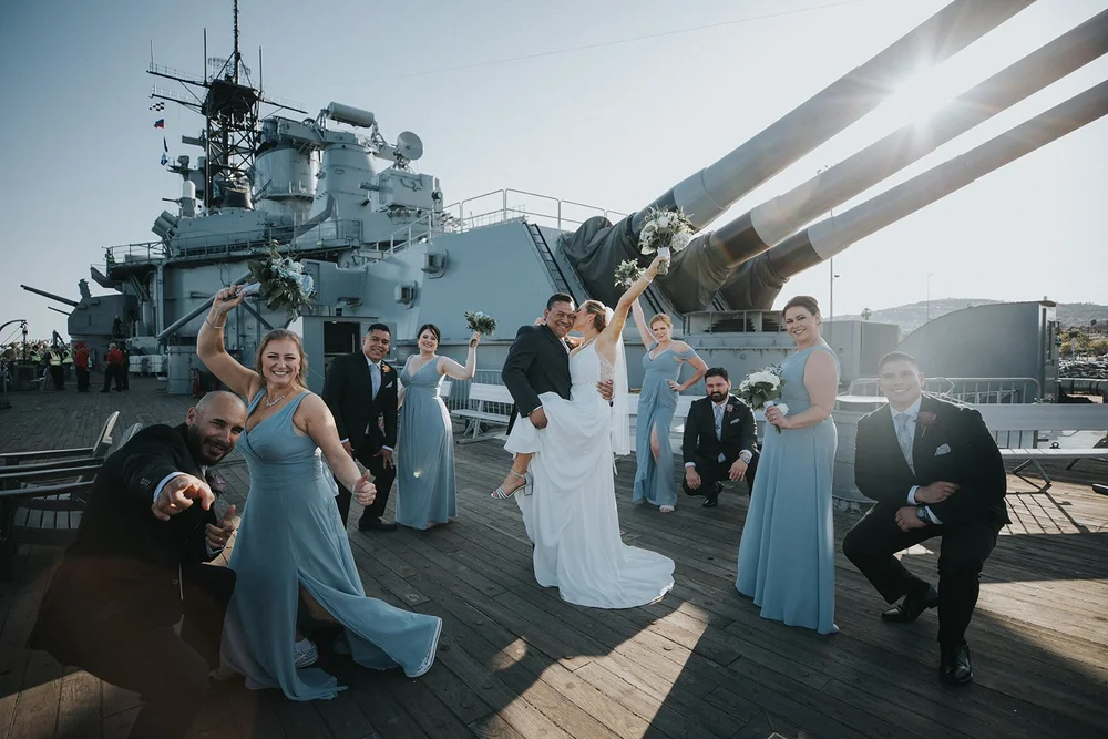 Wedding party celebrating with raised arms as the sun flares through the ship’s guns