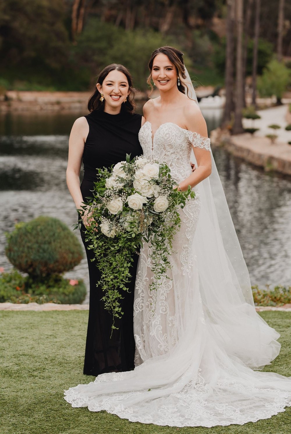 Bride in an off-the-shoulder lace gown with a long-line bodice and sweep train standing on a lawn beside a bridesmaid in black