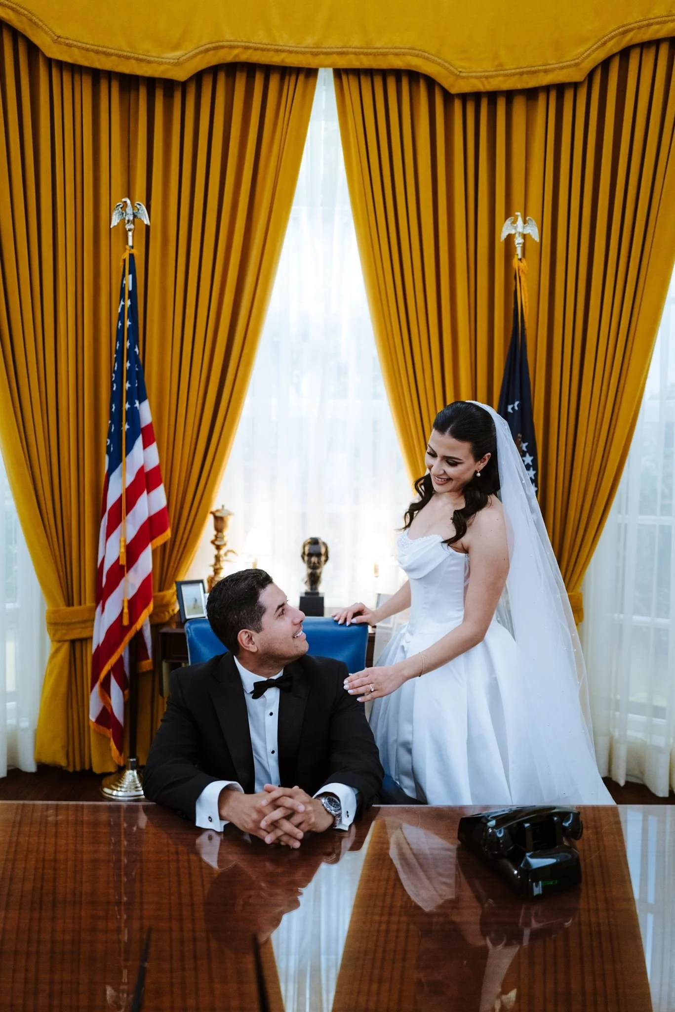 Groom seated at the desk in the Oval Office replica as the bride stands beside him at the Nixon Library
