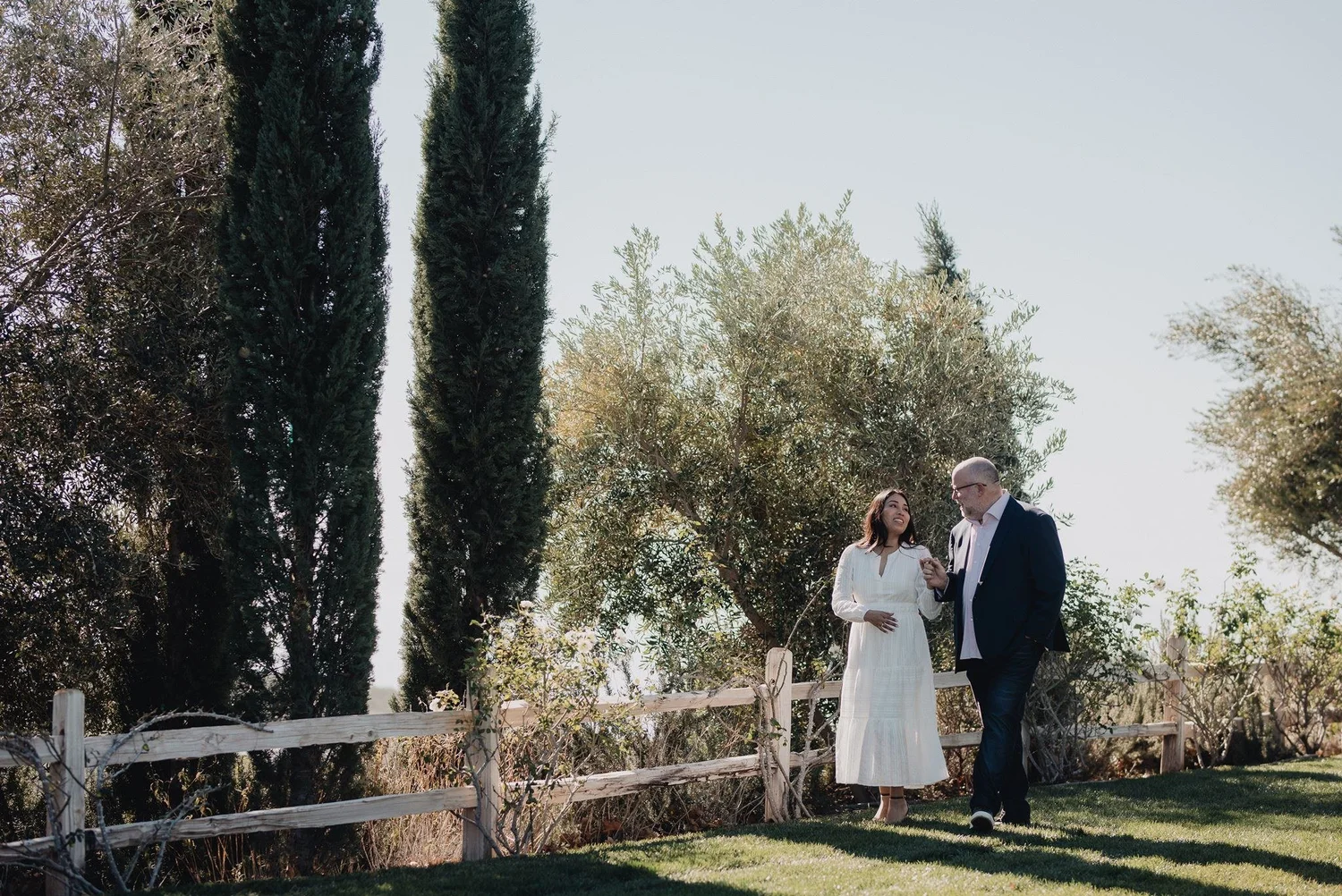 Couple walking past cypress and olive trees by a split-rail fence at a private estate