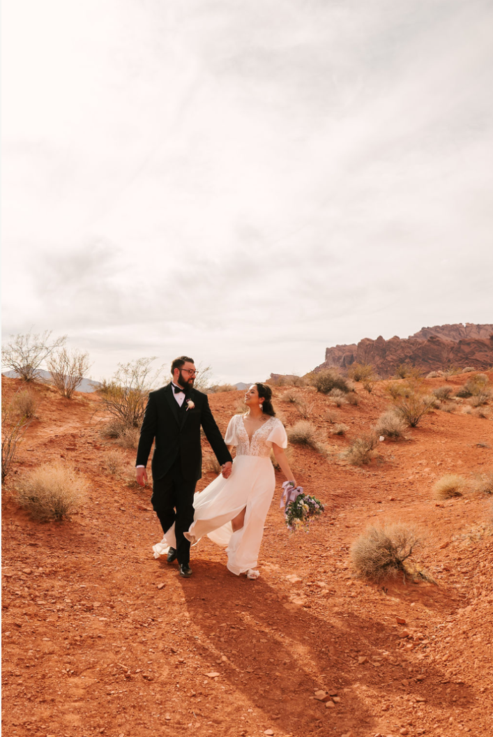 Groom in a tuxedo and bride in a flowing white dress walk hand in hand through red desert scrub at Valley of Fire, her skirt catching the wind.