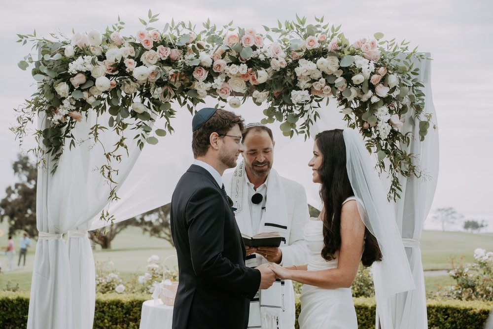 Bride and groom facing each other beneath a floral chuppah while the rabbi reads