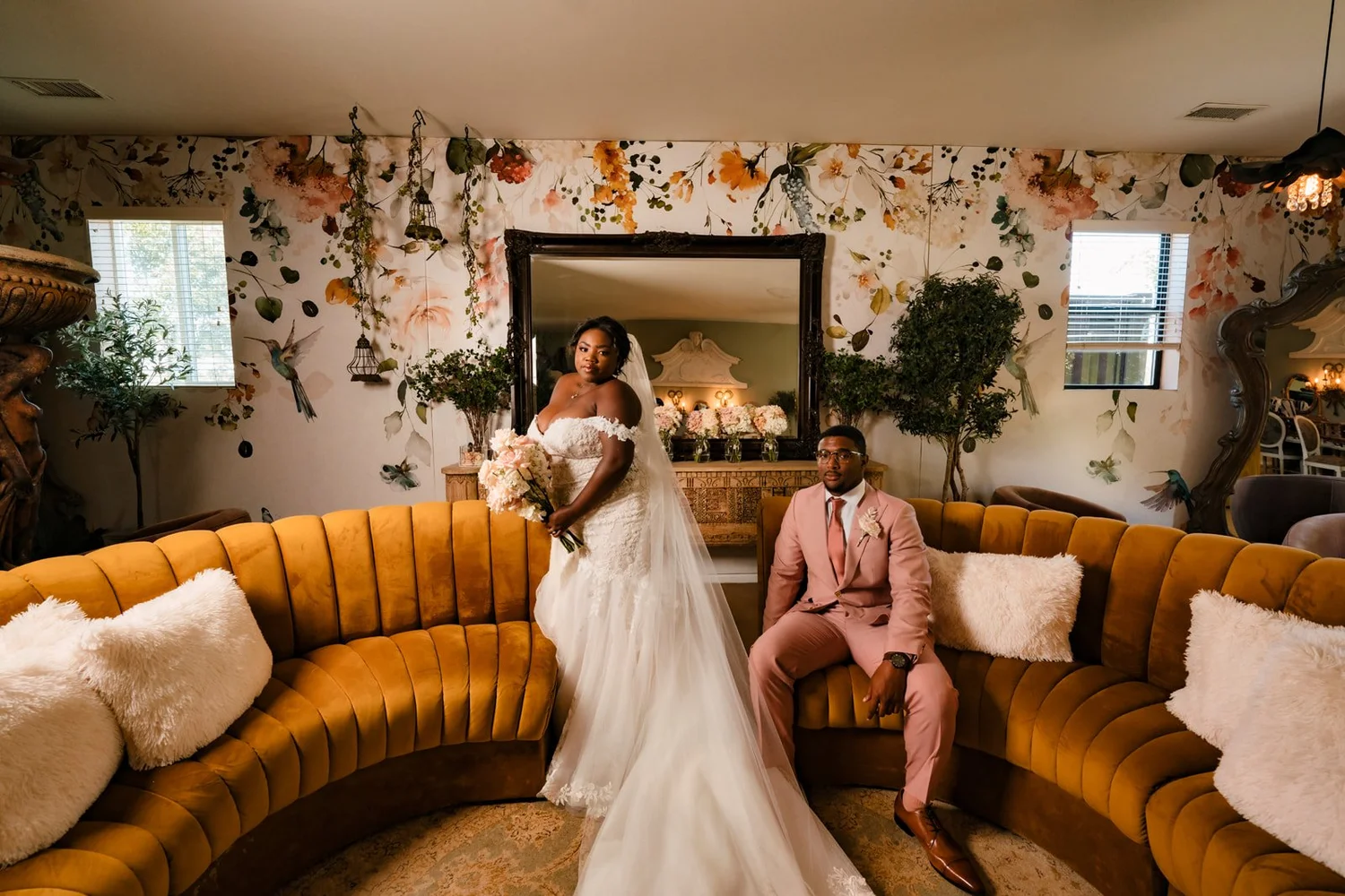 Bride and groom on a curved velvet banquette in the floral-papered lounge at Ethereal Gardens in Escondido