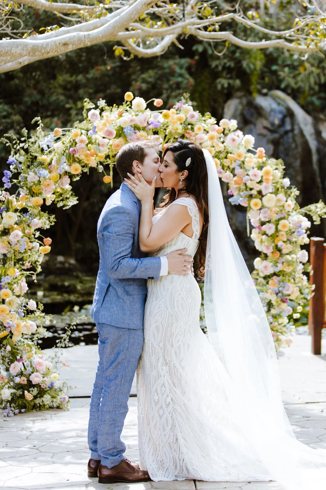 Bride and groom kissing beneath a pastel floral arch at Botanica in Oceanside, dense planting and a pond behind them