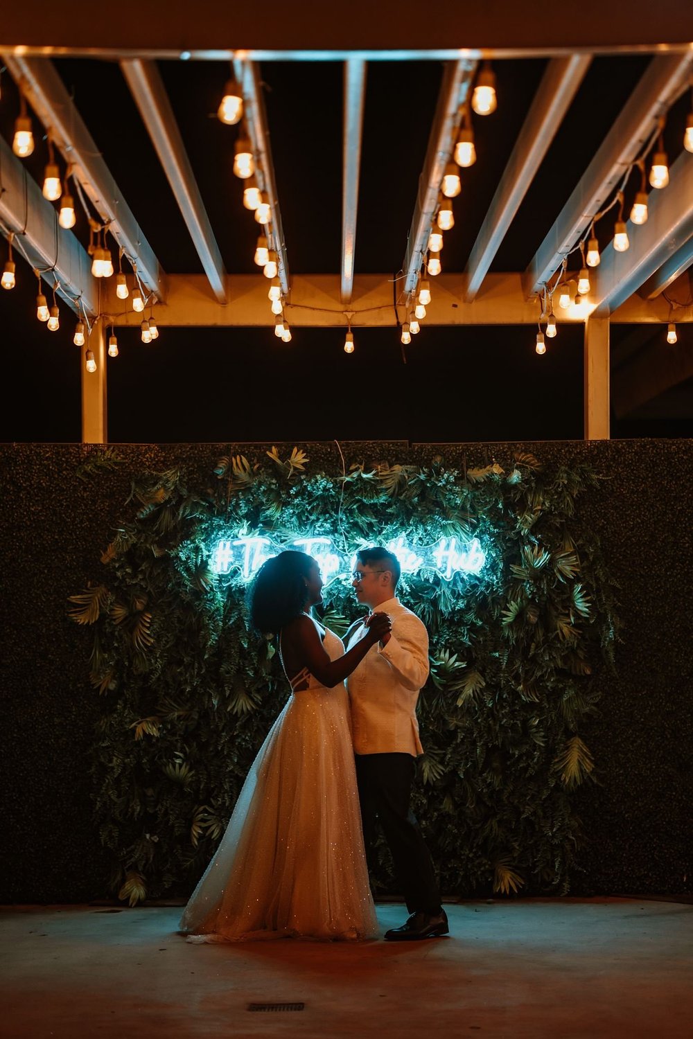 First dance under festoon lights in front of a greenery wall lit by the venue’s cyan neon sign