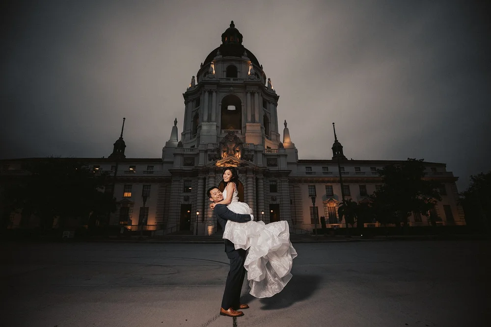 Groom lifting the bride in front of the lit dome of Pasadena City Hall at dusk