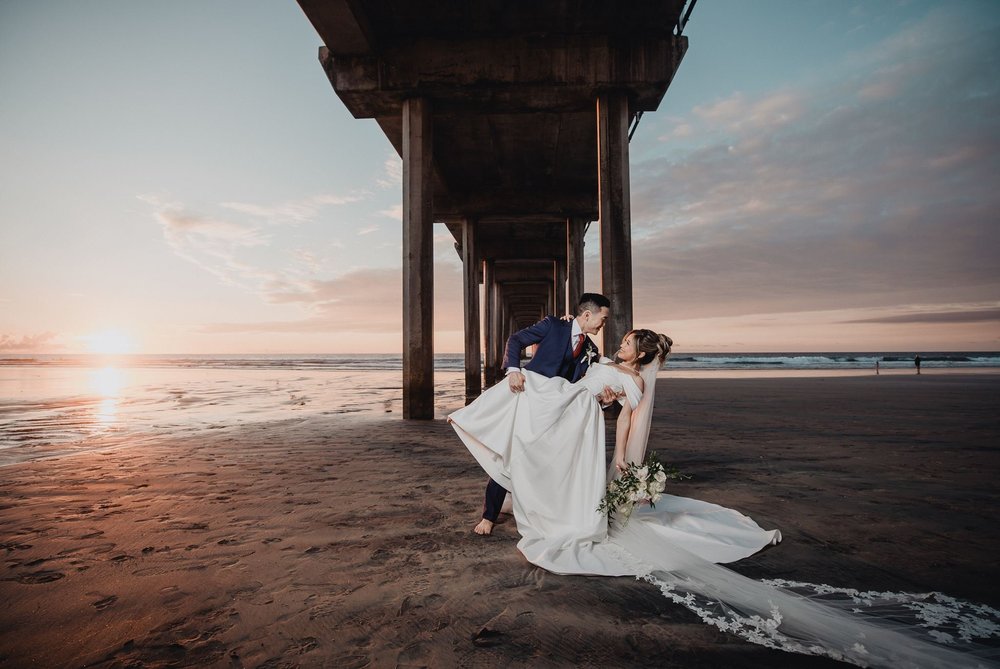 Groom dipping the bride beneath Scripps Pier at sunset